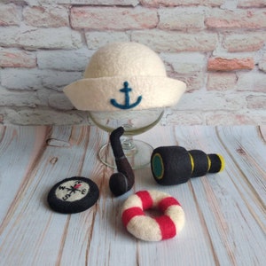 May include: A white felt sailor hat with a blue anchor, a black felt compass, a black felt pipe, a black felt telescope with a green and yellow lens, and a white felt life preserver with red stripes.