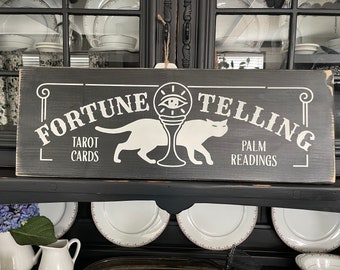 May include: A black wooden sign with white lettering that reads "Fortune Telling" with the words "Tarot Cards" and "Palm Readings" below. The sign features a stylized eye with a cat on either side.