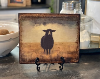 May include: A black sheep standing in a field of tall grass. The sheep is looking directly at the viewer. The background is a blurry image of a cloudy sky. The image is printed on a wooden plaque with a distressed finish.