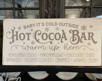 May include: White wooden sign with the text "BABY IT'S COLD OUTSIDE" and "HOT COCOA BAR" in gray cursive lettering. The sign also includes the text "warm up here" and a list of toppings: "PEPPERMINT STICKS MARSHMALLOWS CHOCOLATE CHIPS SPRINKLES CRUSHED COOKIES CARAMELS WHIPPED CREAM".