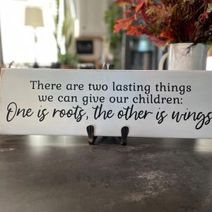 May include: A rectangular, white wooden sign with black text that reads, "There are two lasting things we can give our children: One is roots, the other is wings." The sign is propped up on a black metal stand. A vase of fall foliage is in the background.