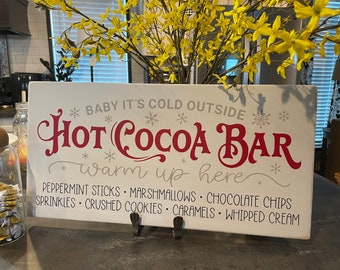 May include: A rectangular white wooden sign with the words "Hot Cocoa Bar" in red script. The sign also includes the text "Baby it's cold outside" and a list of cocoa toppings. Yellow flowers are in the background.