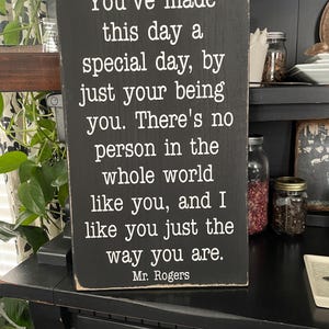 Puede incluir: Un letrero de madera negro con texto blanco. El texto dice: "Has hecho de este d&iacute;a un d&iacute;a especial, simplemente siendo t&uacute;..." Firmado "Mr. Rogers". El letrero se muestra sobre una superficie negra.