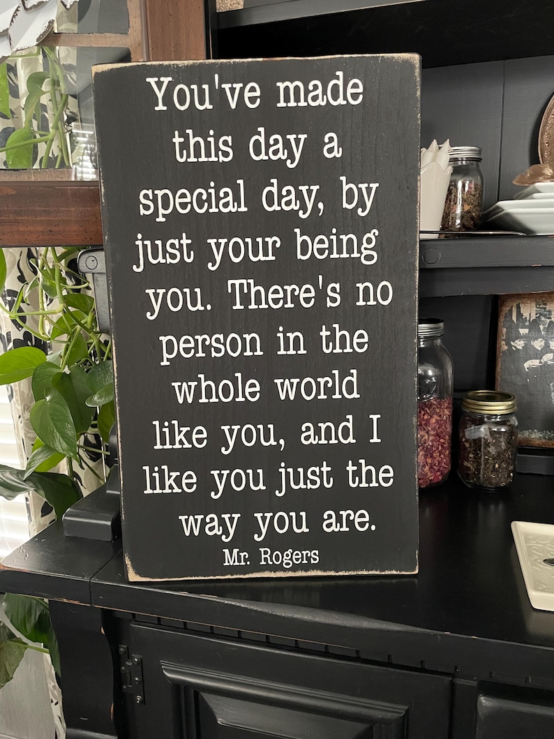 Puede incluir: Un letrero de madera negro con texto blanco. El texto dice: "Has hecho de este d&iacute;a un d&iacute;a especial, simplemente siendo t&uacute;..." Firmado "Mr. Rogers". El letrero se muestra sobre una superficie negra.