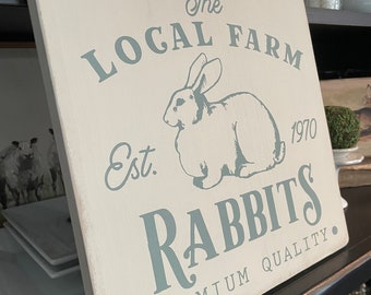 May include: A white wooden sign with the text "The Local Farm Est. 1970 Rabbits Premium Quality." in blue lettering. A white rabbit illustration is in the center of the sign.