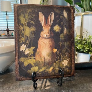 May include: A brown rabbit with long ears sits in a field of green plants and yellow flowers. The rabbit is surrounded by greenery and has a dark brown background. The image is printed on a square piece of wood with a brown frame.
