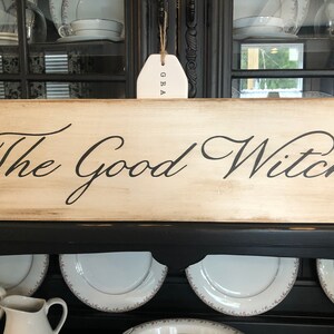 May include: A wooden sign with a distressed white paint finish and black lettering that reads "The Good Witch".