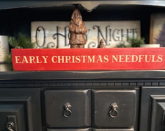 May include: A red wooden sign with the words "EARLY CHRISTMAS NEEDFULS" in gold lettering. The sign is displayed on a black cabinet. A Santa Claus figurine and a sign that says "O Holy Night" are in the background.