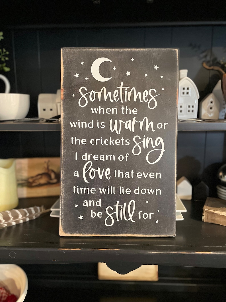 Op de afbeelding: Een zwart houten bord met witte tekst en een halve maan. De tekst luidt "Sometimes when the wind is warm or the crickets sing I dream of a love that even time will lie down and be still for."