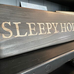 Puede incluir: Un letrero de madera gris oscuro con las palabras "SLEEPY HOLLOW" pintadas en blanco.
