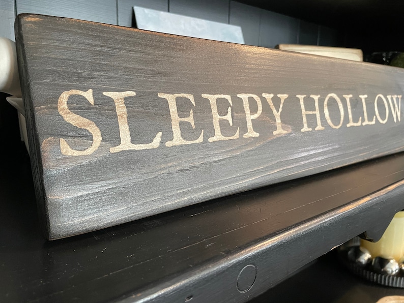 Puede incluir: Un letrero de madera gris oscuro con las palabras "SLEEPY HOLLOW" pintadas en blanco.