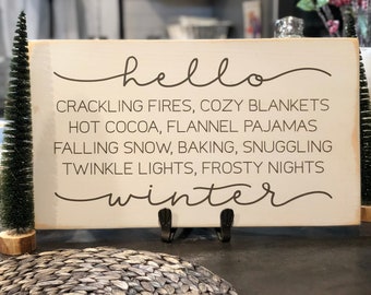 May include: White wooden sign with brown text that reads "hello winter" and lists winter activities like crackling fires, cozy blankets, hot cocoa, flannel pajamas, falling snow, baking, snuggling, twinkle lights, and frosty nights.
