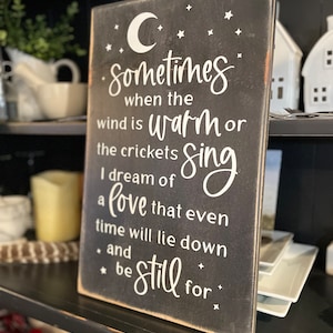 Op de afbeelding: Een zwart houten bord met witte tekst die luidt: "Sometimes when the wind is warm or the crickets sing I dream of a love that even time will lie down and be still for."