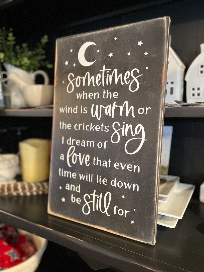 Op de afbeelding: Een zwart houten bord met witte tekst die luidt: "Sometimes when the wind is warm or the crickets sing I dream of a love that even time will lie down and be still for."