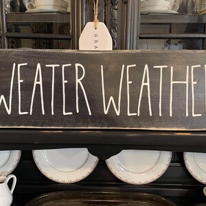 May include: A black wooden sign with white painted lettering that reads "SWEATER WEATHER".