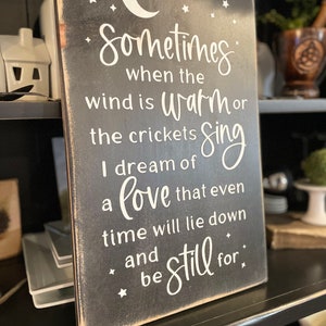 Op de afbeelding: Een zwart houten bord met witte tekst die luidt: "Sometimes when the wind is warm or the crickets sing I dream of a love that even time will lie down and be still for."