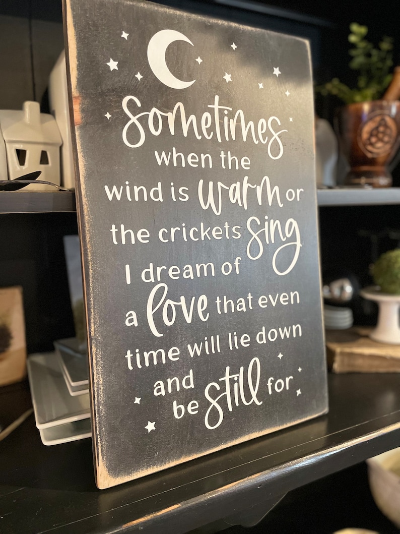 Op de afbeelding: Een zwart houten bord met witte tekst die luidt: "Sometimes when the wind is warm or the crickets sing I dream of a love that even time will lie down and be still for."