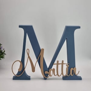 Op de afbeelding: Een grote, blauwe houten letter "M" met de naam "Mattia" in een lichter bruin script. De letter is vrijstaand en waarschijnlijk bedoeld voor woondecoratie of een persoonlijk cadeau. De achtergrond is effen wit.