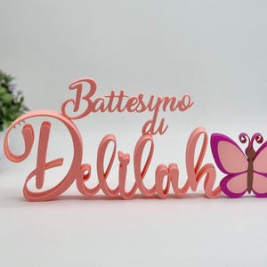 Può includere: Decorazione per battesimo con il nome "Delilah" in corsivo, accompagnato da una farfalla rosa e viola. Sono presenti anche le parole "Battesimo di". Ideale per una celebrazione battesimale.