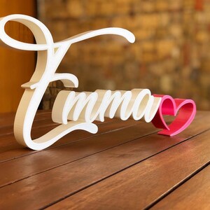 Può includere: Un'insegna tridimensionale bianca che riporta il nome Emma in un carattere corsivo. Le lettere hanno una finitura liscia e sono posizionate in verticale. Un piccolo cuore rosa pieno è fissato sul lato destro del nome.