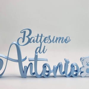 Può includere: Un'insegna in legno azzurro con le parole "Battesimo di Antonio" in corsivo. Una piccola figura di orsetto è sulla destra. L'insegna è un oggetto decorativo per un battesimo.