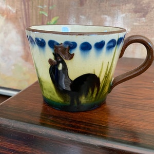 May include: Hand-painted ceramic mug with a rooster design. The mug features a cream-colored base with a dark brown handle and rim. The rooster is black with a brown comb, set against green and blue accents.