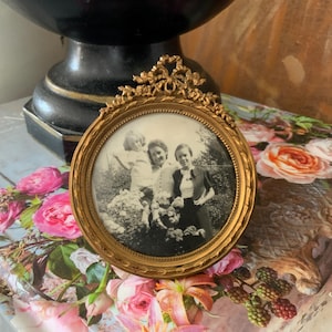 May include: A vintage, round, gold-toned picture frame with an ornate top detail. The frame holds a black and white photograph of three people in a garden setting. The frame rests on a floral book and is surrounded by pink flowers.