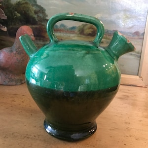 May include: A two-toned green ceramic pitcher with a handle and two spouts. The upper portion is a bright green, while the lower half is a darker shade. The pitcher has a rounded body and a pedestal base. It is likely an antique or vintage item.