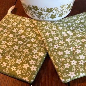 May include: Two green quilted pot holders with a white floral pattern. The pot holders have a white border and a loop for hanging.