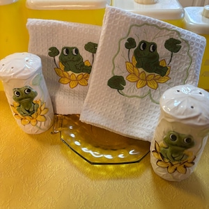 May include: A collection of kitchen items featuring a frog theme. Includes two white salt shakers with frog and flower designs, two white waffle-weave towels with frog embroidery, and a yellow glass plate. Yellow canisters are in the background.
