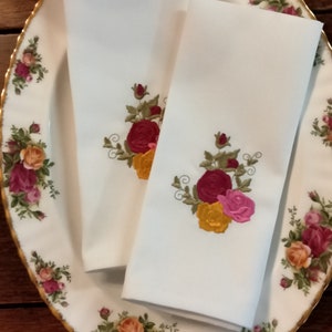 May include: Two white linen napkins with embroidered floral designs. The napkins feature a bouquet of red, yellow, and pink roses with green leaves.
