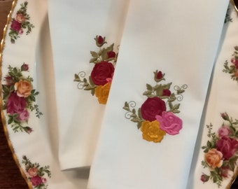 May include: Two white linen napkins with embroidered floral designs. The napkins feature a bouquet of red, yellow, and pink roses with green leaves.