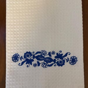 May include: White waffle-weave kitchen towel with a blue floral embroidery design. The embroidered border features stylized flowers and leaves. The towel is rectangular and appears to be made of cotton.