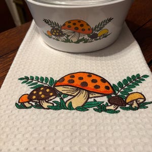 Könnte beinhalten: Eine weiße Auflaufform aus Keramik mit einem klaren Glasdeckel und einem Pilz- und Farnmotiv, kombiniert mit einem weißen Waffelpique-Küchentuch, das mit einem passenden Design aus orangefarbenen und braunen Pilzen und grünen Farnen bestickt ist.