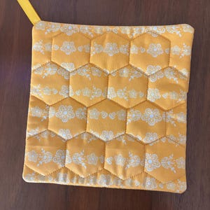 May include: A square, quilted pot holder in a honeycomb pattern. The fabric is a golden yellow with a white floral design. A yellow ribbon loop is attached to one corner for hanging. The pot holder is approximately 8 inches square.