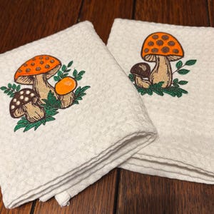 May include: Two white waffle-weave kitchen towels featuring embroidered mushroom designs. One towel displays two mushrooms with orange and brown caps, while the other shows a single mushroom with an orange cap, all surrounded by green leaves.
