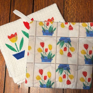 May include: A square quilted pot holder featuring a repeating pattern of red and yellow tulips in blue pots, paired with a white waffle-weave kitchen towel embroidered with a single red and yellow tulip in a blue pot and a white hanging loop.