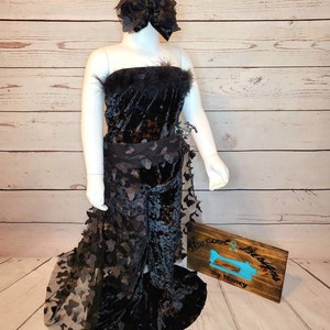 May include: A black velvet dress with a black tulle overlay and butterfly cutouts. The dress has a strapless bodice and a fitted silhouette. The dress is accessorized with a black flower headband. The dress is displayed on a mannequin against a woodgrain background. The sign in the image reads "Sew Cool Designs by Nancy".