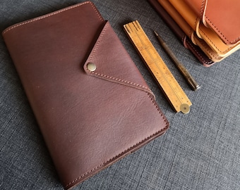 May include: Three brown leather journals with a flap closure and a metal button. The journals are stacked on top of each other. A wooden ruler and a metal pen are lying on a gray surface.