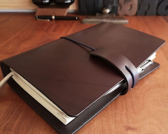 May include: A dark brown leather bound journal with a pen holder and a strap closure. The journal is closed and lying on a wooden surface.