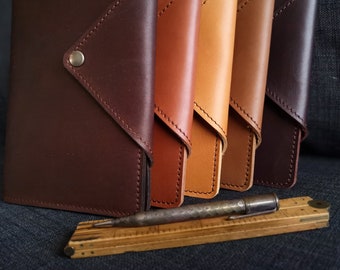 May include: Five leather journals in different shades of brown, from dark brown to light tan, lined up in a row. The journals have a flap closure and a silver button.
