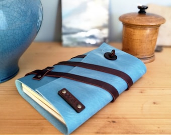 May include: A blue leather journal with brown leather straps and a metal closure. The journal is closed and lying on a wooden surface.