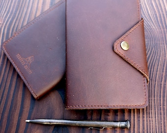 May include: Two brown leather wallets with a pen on a wooden surface. The wallet on the left has the text "FEATHER RIVER" embossed on the front. The wallet on the right has a flap with a gold button.