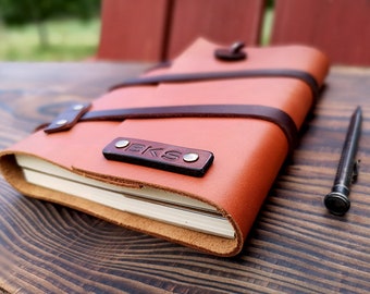 May include: A brown leather journal with a brown leather strap and a metal buckle. The journal has the initials "BKS" embossed on the front cover.