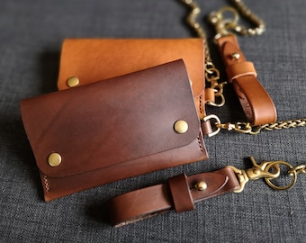 May include: Two leather wallets, one brown and one tan, connected by a gold chain. The wallets have brass accents and are made of high-quality leather.