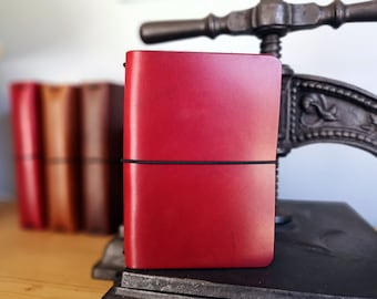 May include: A red leather notebook with a black elastic band closure. The notebook is closed and sitting on a metal surface.