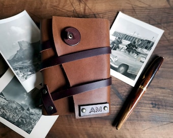 May include: A brown leather journal with a dark brown strap closure and a metal plate with the initials "AM" on the front. The journal is sitting on a wooden surface with two black and white photographs and a brown pen.