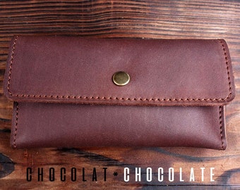 May include: A brown leather wallet with a flap closure and a brass button. The wallet is on a wooden surface.