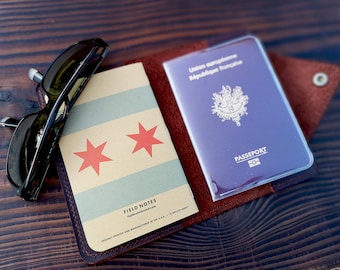 May include: A brown leather passport holder with a French passport inside. The passport holder has a pocket for a notebook, which is visible in the image. The notebook has a Chicago flag design on the cover and the text "FIELD NOTES" printed on the bottom. A pair of sunglasses is lying on the table next to the holder.