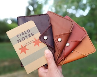 May include: Five leather notebook covers in different shades of brown, from dark brown to light tan. The covers are folded in half and have a snap closure. A small Field Notes memo book is in front of the covers.
